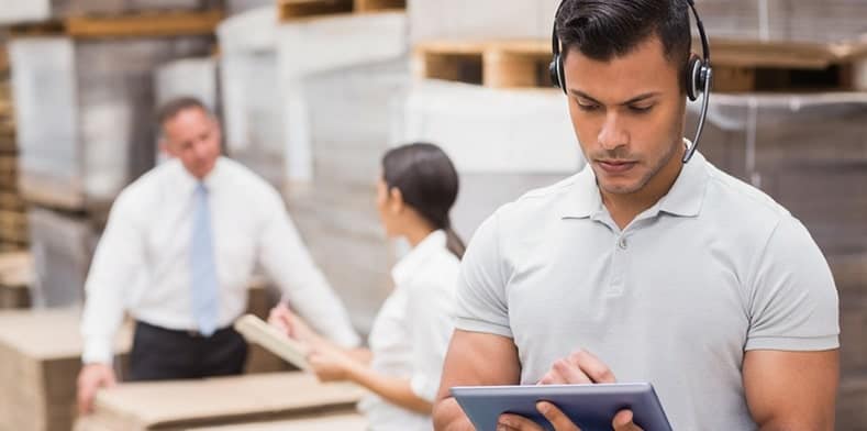 Man on tablet with headset on in warehouse with staff in background