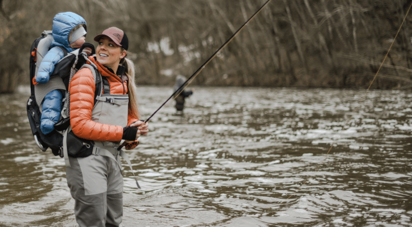 Person flyfishing in river with baby starpped to back