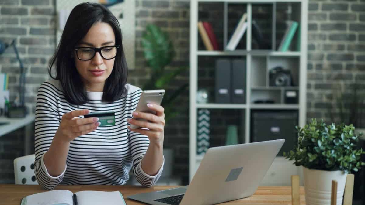 Woman at a desk using a smartphone and credit card to make an online purchase.