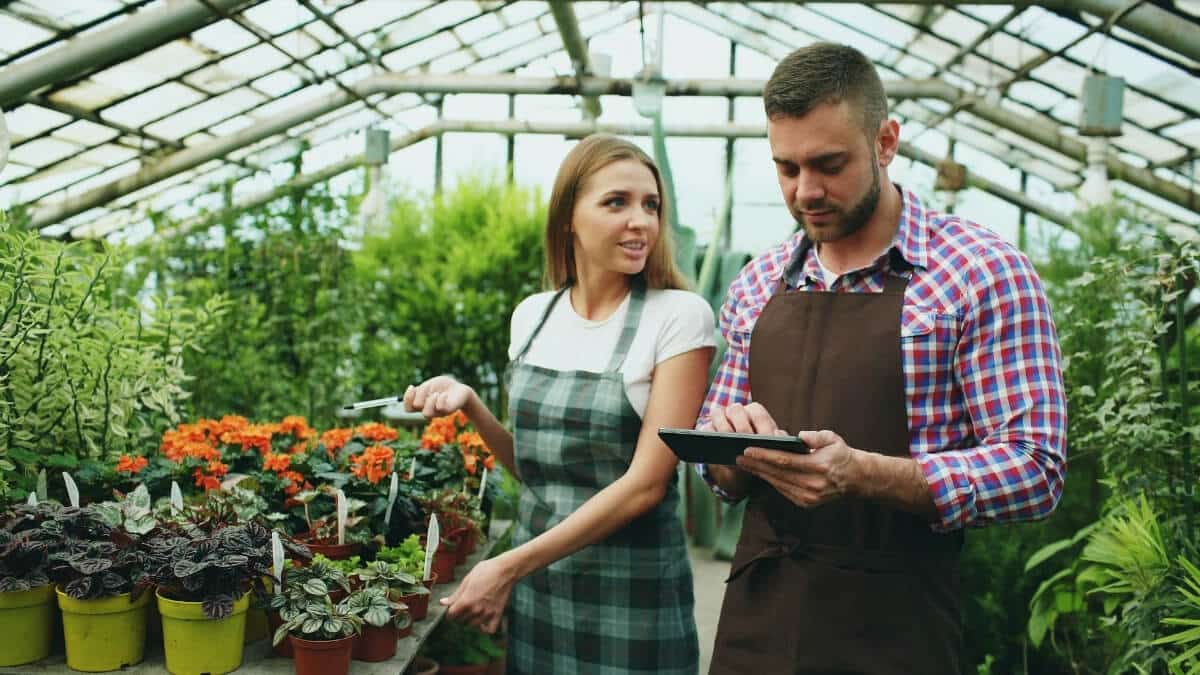 A male and female warehouse manager using a digital tablet to track and manage plant inventory inside a large commercial greenhouse.